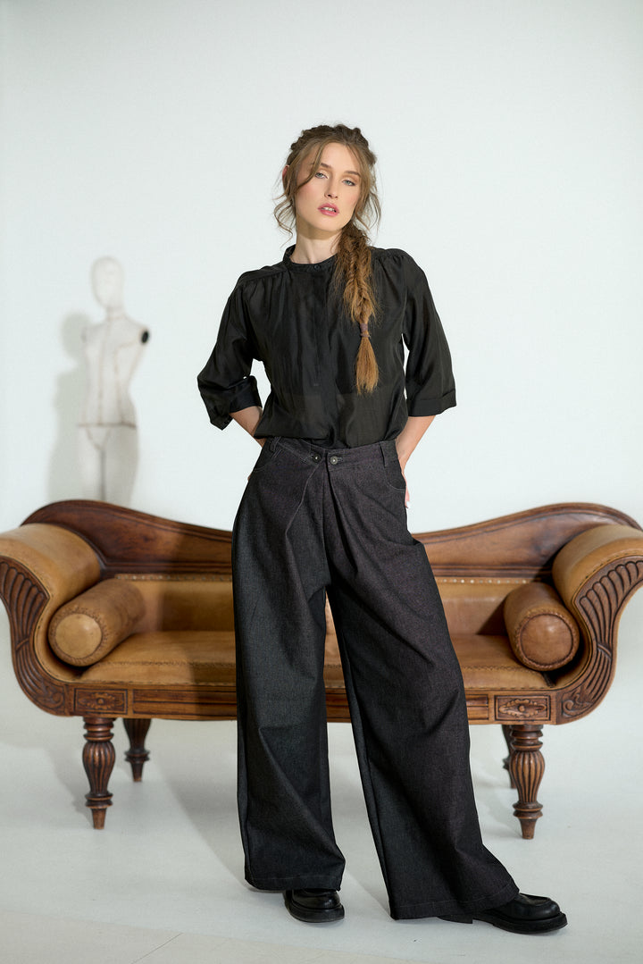 A woman stands in a studio wearing a black long-sleeve blouse and dark wide-leg trousers, posed in front of a brown wooden bench against a white background.