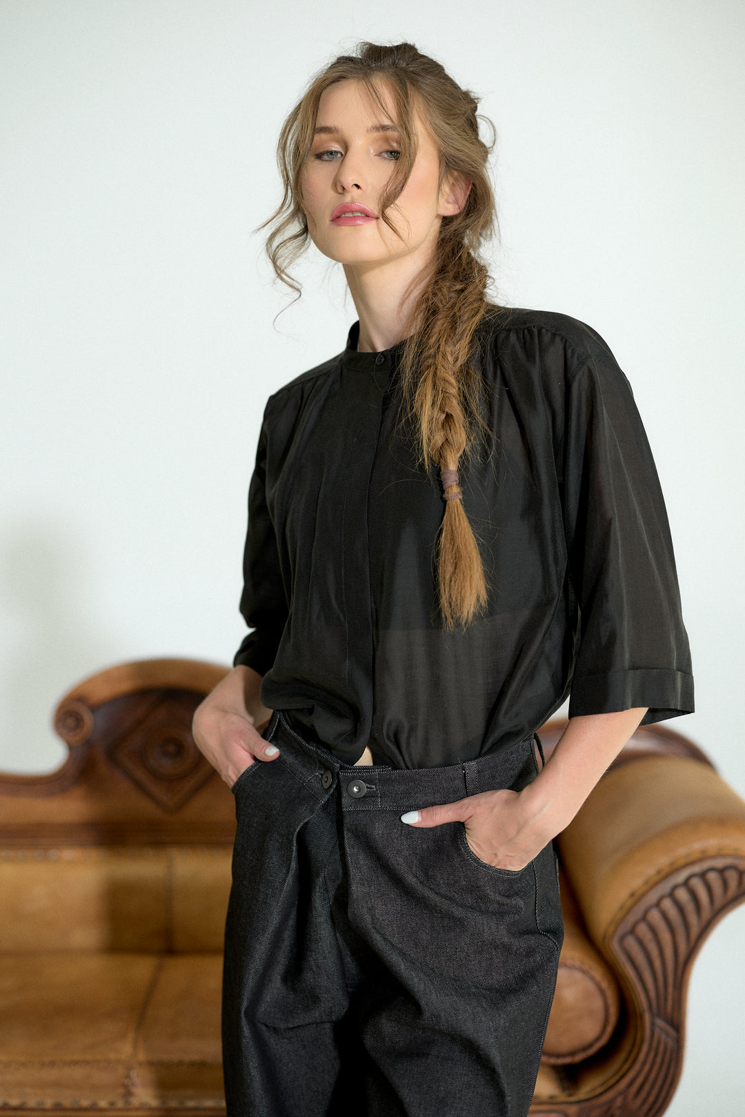 A woman with a long blonde braid wearing a black short-sleeve top and dark charcoal trousers, standing in front of a brown leather sofa against a white wall.