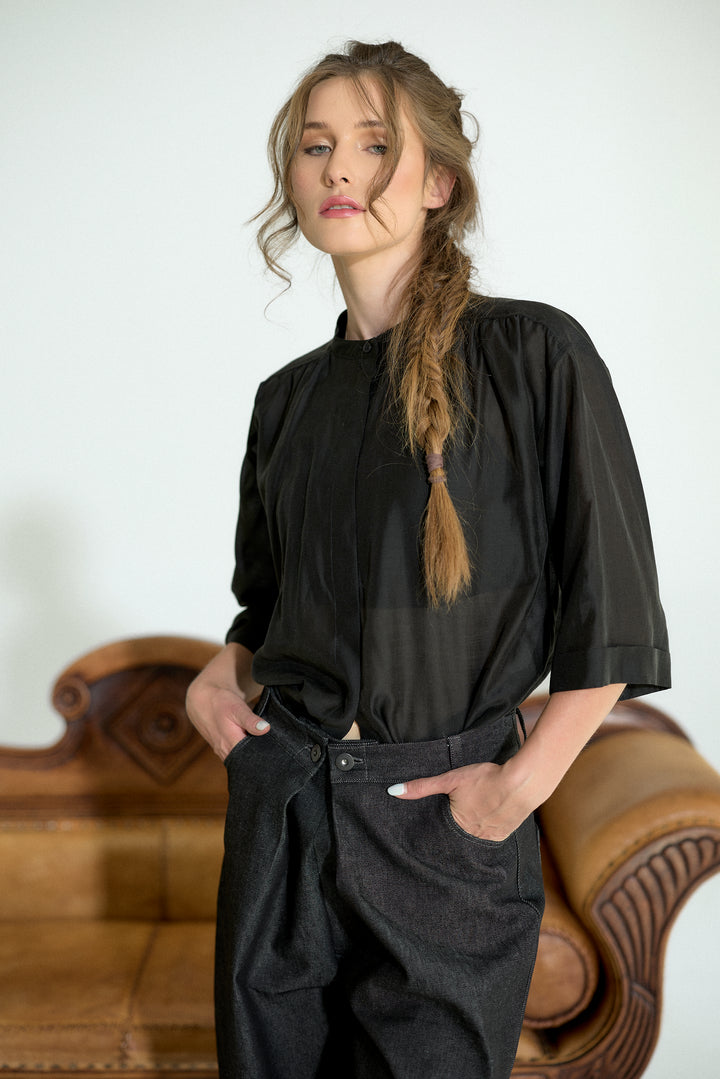 A woman with a long blonde braid wearing a black short-sleeve top and dark charcoal trousers, standing in front of a brown leather sofa against a white wall.