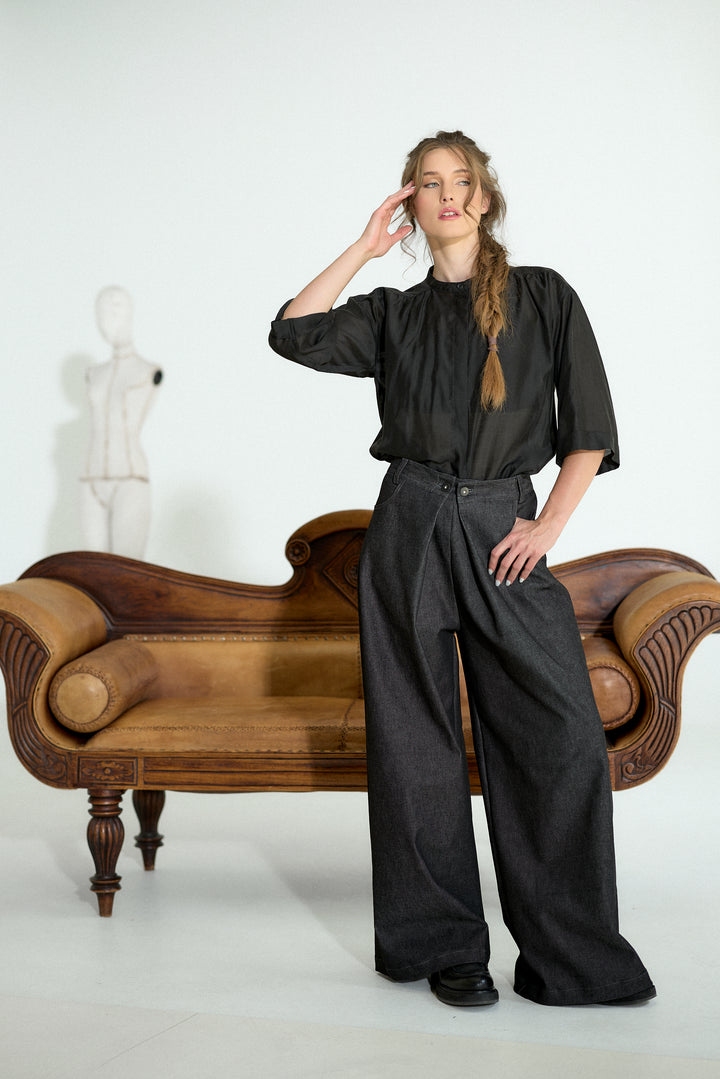A model stands in a studio setting wearing a black short-sleeve blouse and wide-leg charcoal gray trousers, posed next to a brown leather chaise lounge.