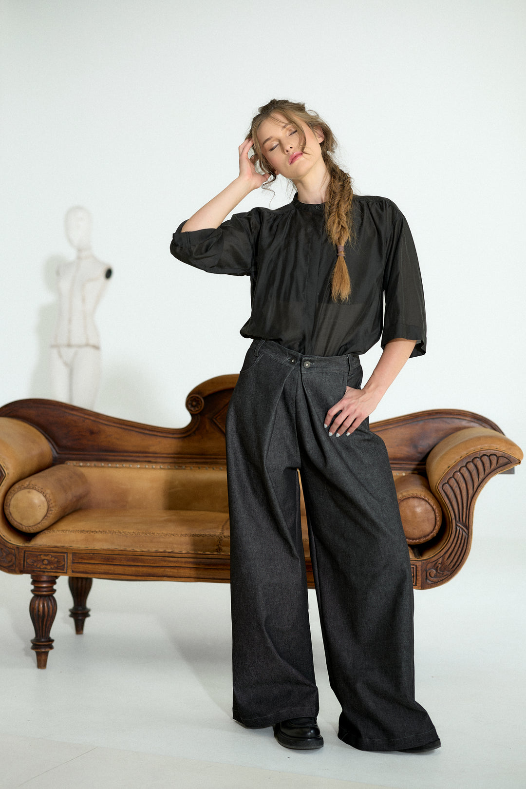 A woman posing in a black short-sleeve blouse and dark gray wide-leg trousers, standing in front of a vintage wooden sofa against a white studio background.