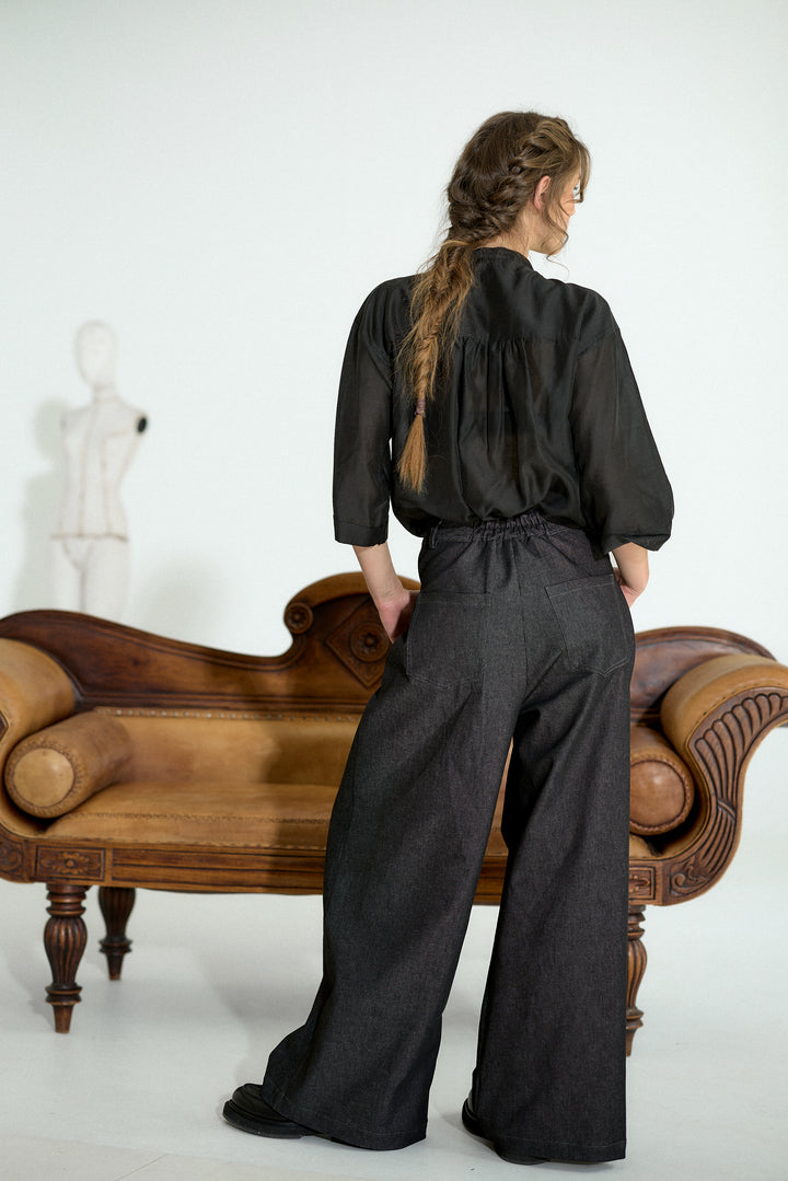 A woman stands with her back to the camera wearing a black long-sleeve top and wide-leg dark trousers, posed in front of a brown wooden chaise lounge against a white studio background.