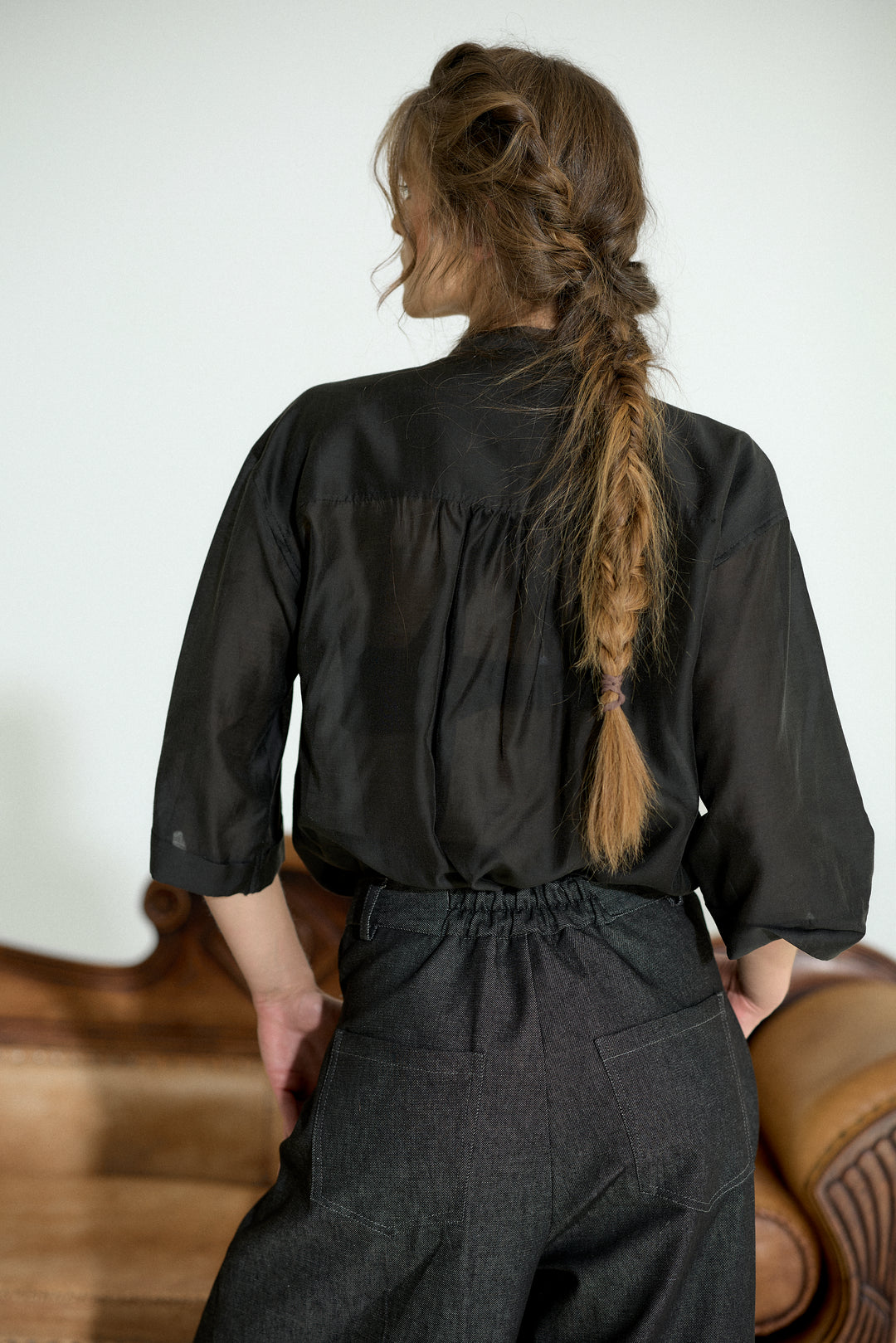 A woman shown from behind wearing a black three-quarter sleeve blouse and dark charcoal wide-leg trousers, standing in an indoor setting with a light-colored wall and wooden furniture visible.