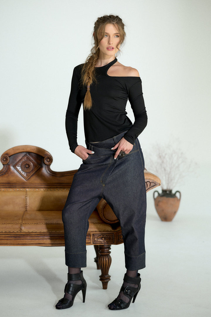 A woman posing in a studio setting wearing a black asymmetrical long-sleeve top and dark gray baggy pants, standing next to a vintage wooden bench.