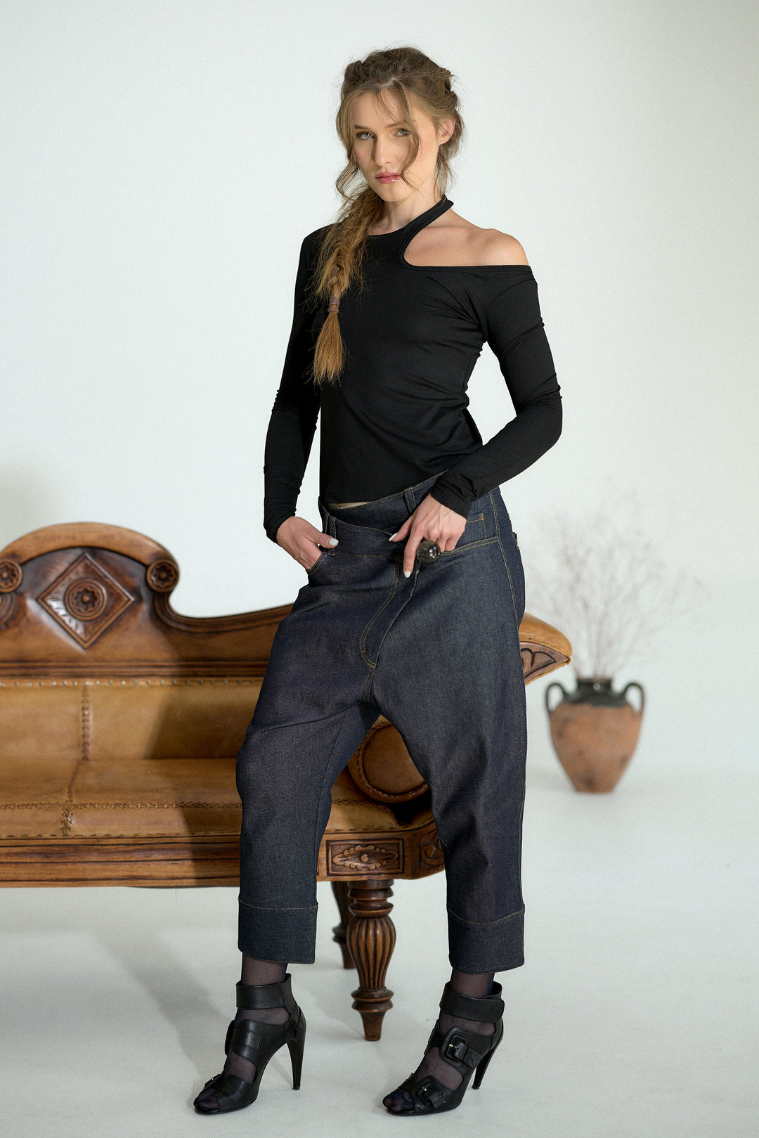 A woman posing in a studio setting, wearing a black asymmetrical off-the-shoulder long-sleeve top, dark gray baggy jeans, and black high-heeled shoes, sitting on a vintage wooden bench.