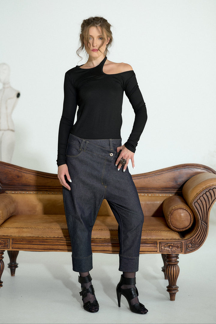 A woman stands in a studio wearing a black asymmetrical long-sleeve top and dark gray baggy cropped pants, posed in front of a brown leather bench.