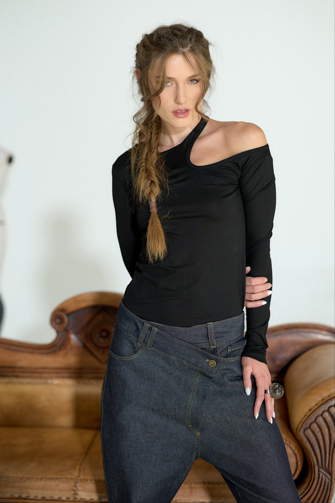 A woman posing in a black off-the-shoulder long-sleeve top and dark gray jeans, standing in front of a light-colored wall and a brown leather sofa.