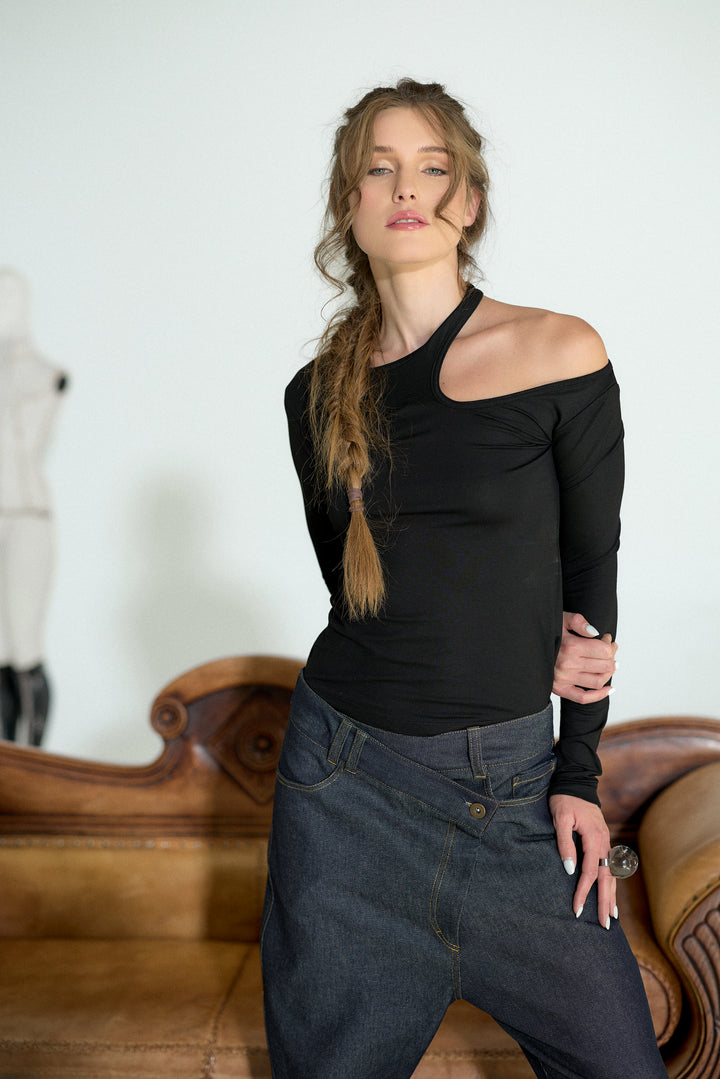 A woman posing in a black off-the-shoulder long-sleeve top and dark blue jeans in front of a light-colored wall and a brown leather sofa.
