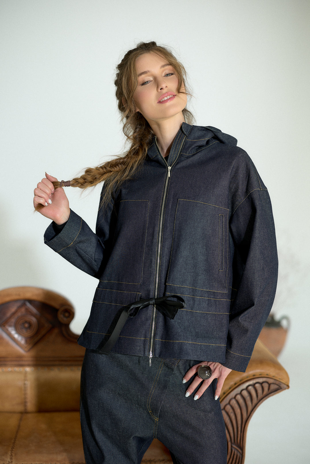 A woman wearing a dark indigo denim hooded jacket with a front zipper, contrast stitching, and a black tie belt at the waist, paired with matching dark denim pants, posed in front of a light-colored wall and a brown leather chair.
