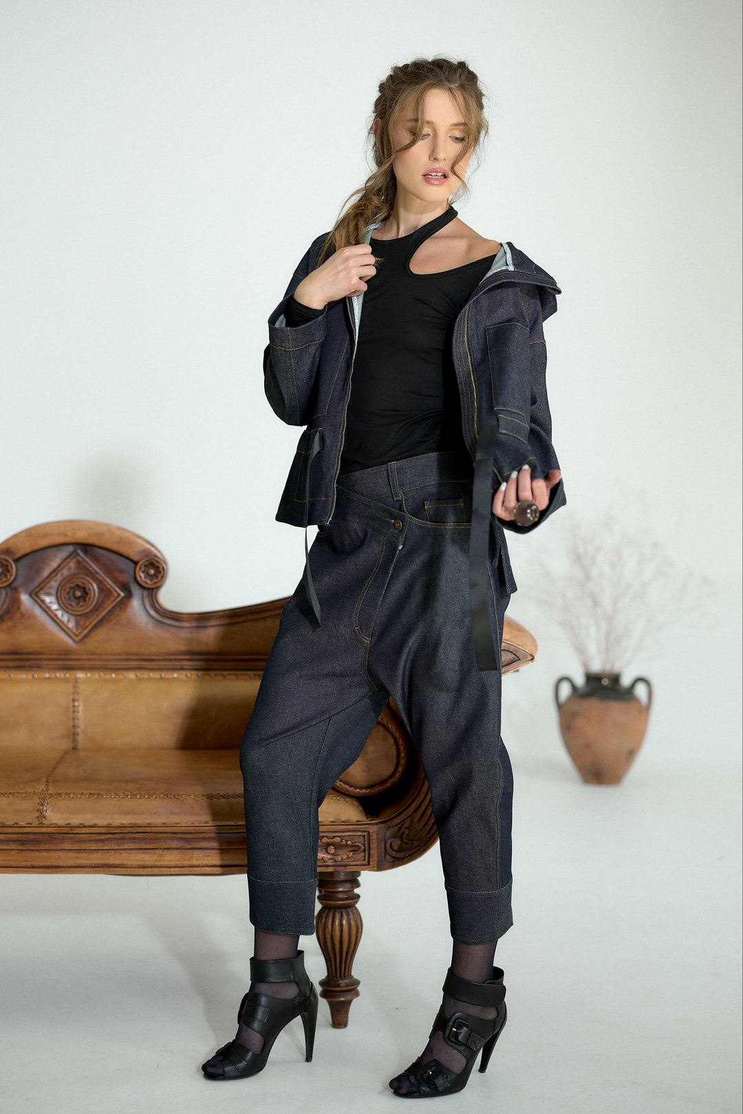 A model wearing a dark denim jacket, dark baggy jeans, a black top, and black high-heeled boots, posing next to a vintage wooden bench in a studio setting.