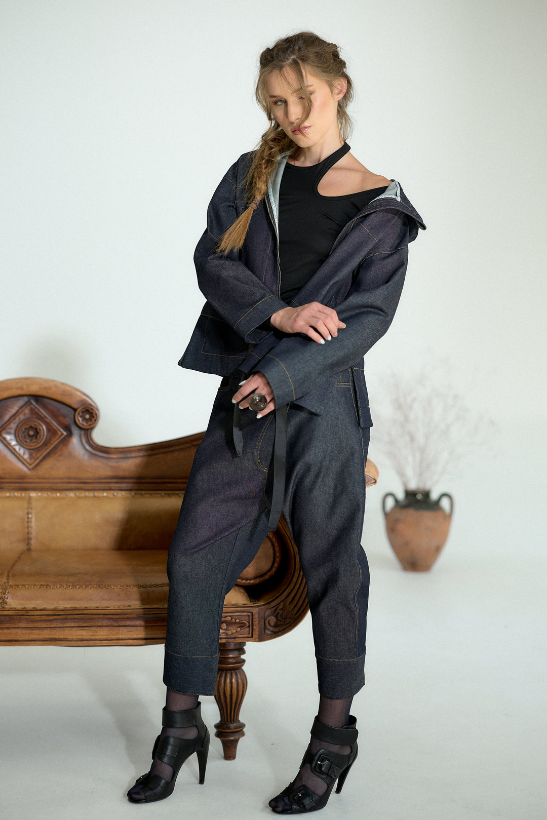 A model wearing a dark gray oversized zip-up jacket, matching wide-leg pants, a black cutout top, and black high-heeled boots, posing next to a vintage wooden bench in a studio setting.