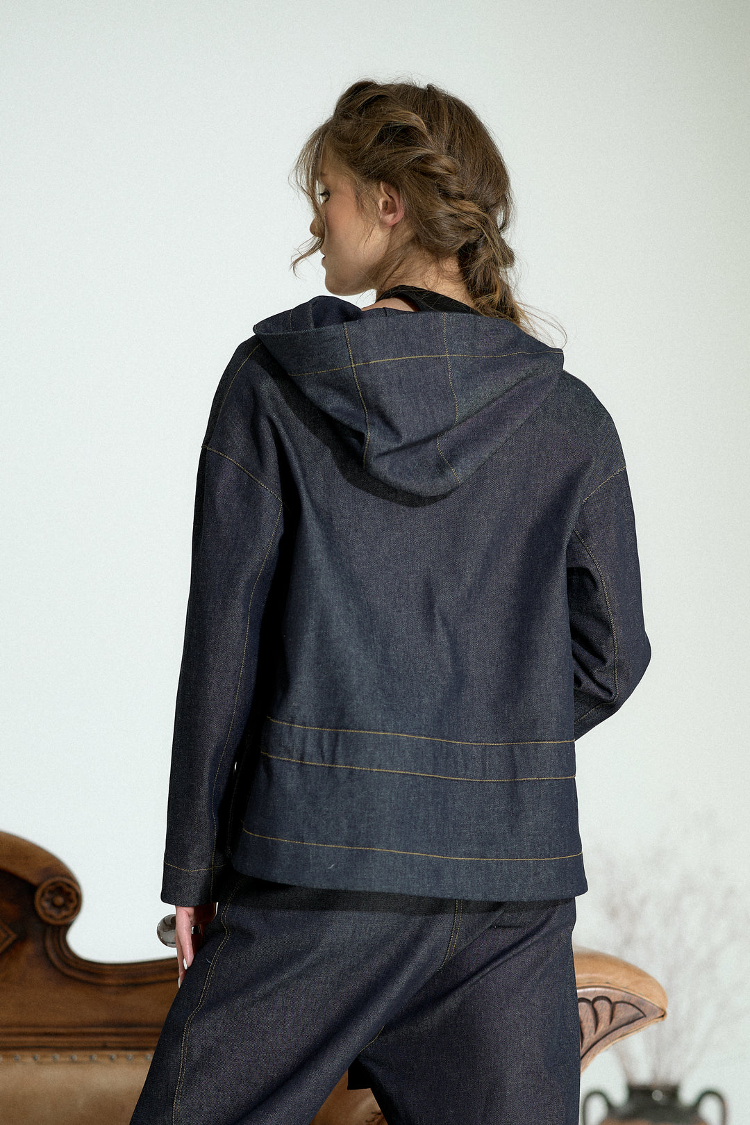 A woman shown from the back wearing a dark indigo denim hooded jacket with contrasting light tan stitching, paired with matching dark denim pants, set against a light gray wall with a brown leather chair partially visible.