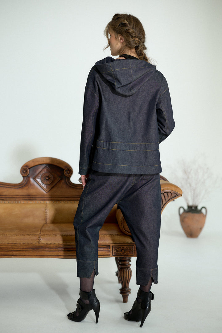 A woman stands with her back to the camera wearing a dark indigo denim hooded jacket and matching cropped trousers, styled with black ankle boots. She is posed in front of a vintage wooden bench in a minimalist studio setting.