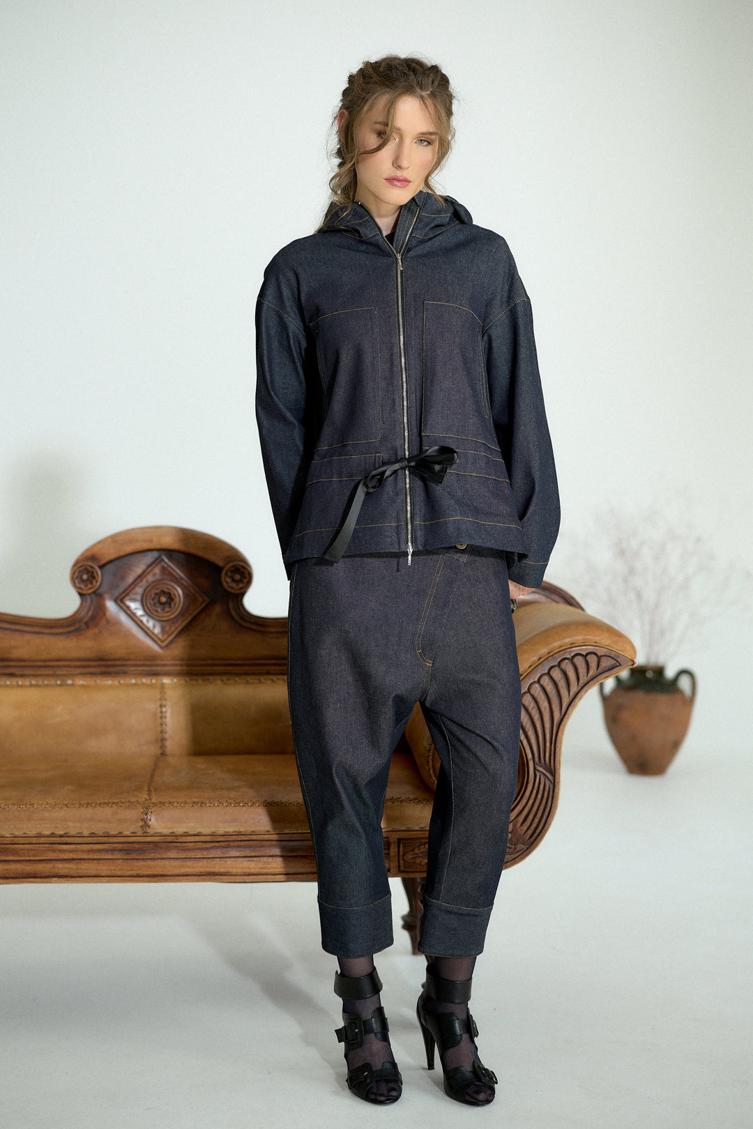 A model stands in a studio wearing a dark indigo denim two-piece set consisting of a zip-up hooded jacket and matching cropped pants. She is posed in front of a vintage wooden bench with a potted plant in the background.