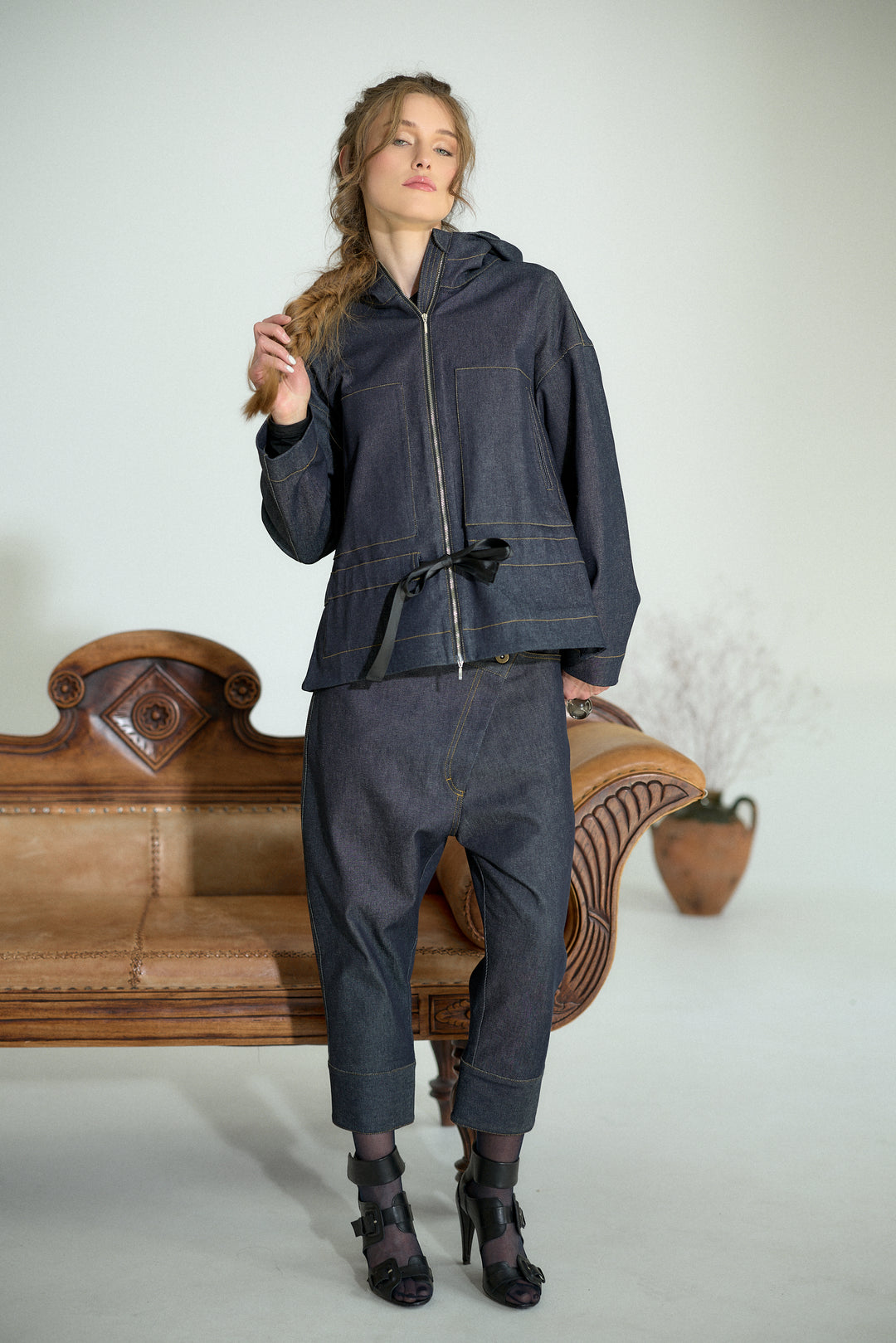 A model wearing a dark indigo denim two-piece set consisting of a hooded zip-up jacket and matching cropped pants, standing in front of a vintage wooden bench.