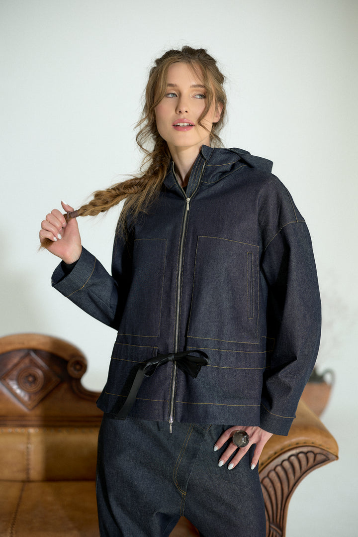 A woman wearing a dark indigo denim hooded jacket with a front zipper, drawstring waist, and contrast stitching, standing in front of a light wall with a wooden chair partially visible.