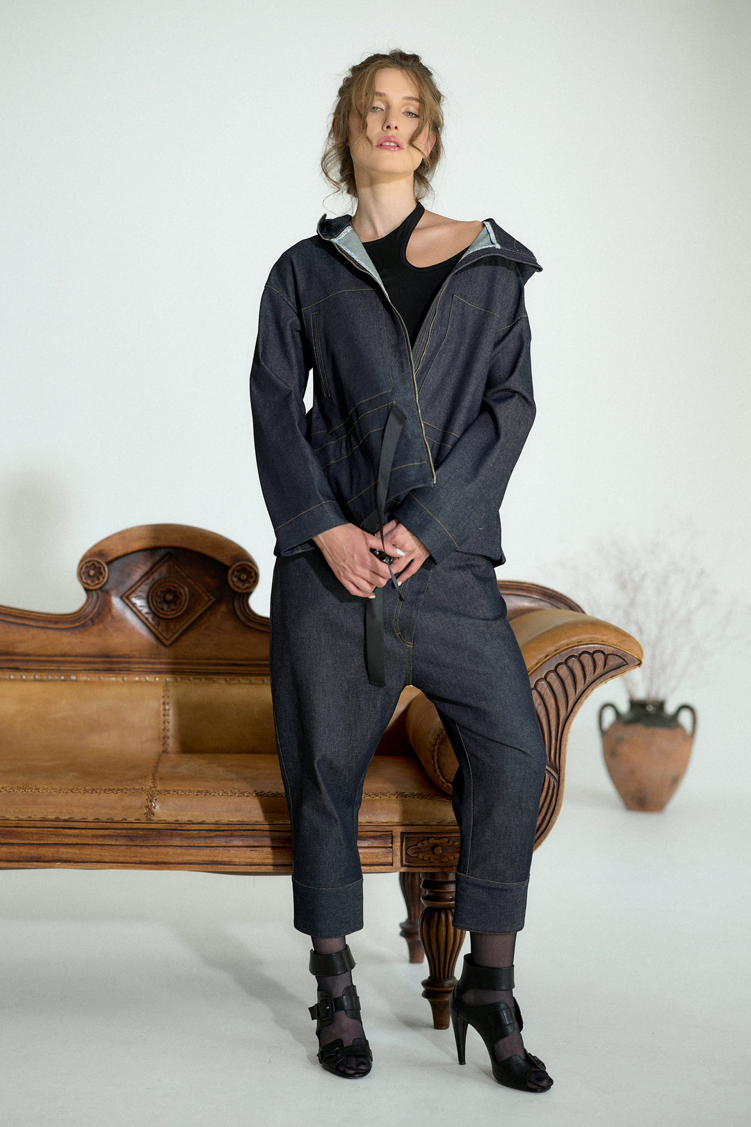A model wearing a dark navy blue hooded zip-up jacket and matching cropped pants, styled with a black top and black high-heeled boots, posing on a vintage brown leather sofa against a white studio background.