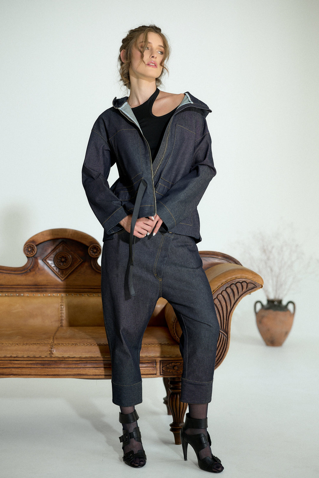 A model wearing a dark indigo denim zip-up hoodie and matching cropped trousers, sitting on an ornate wooden bench against a plain white studio background.