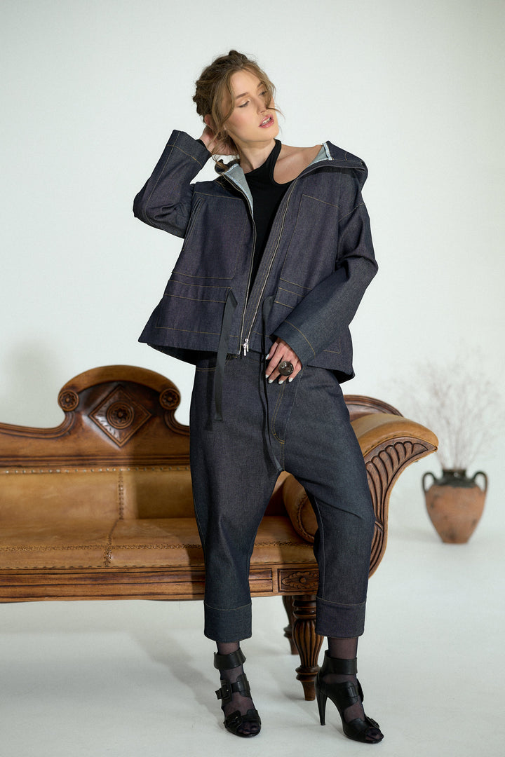 A model wearing a dark indigo denim hooded jacket and matching wide-leg pants, styled with a black top and black high-heeled sandals, posing on a vintage wooden bench against a white studio background.