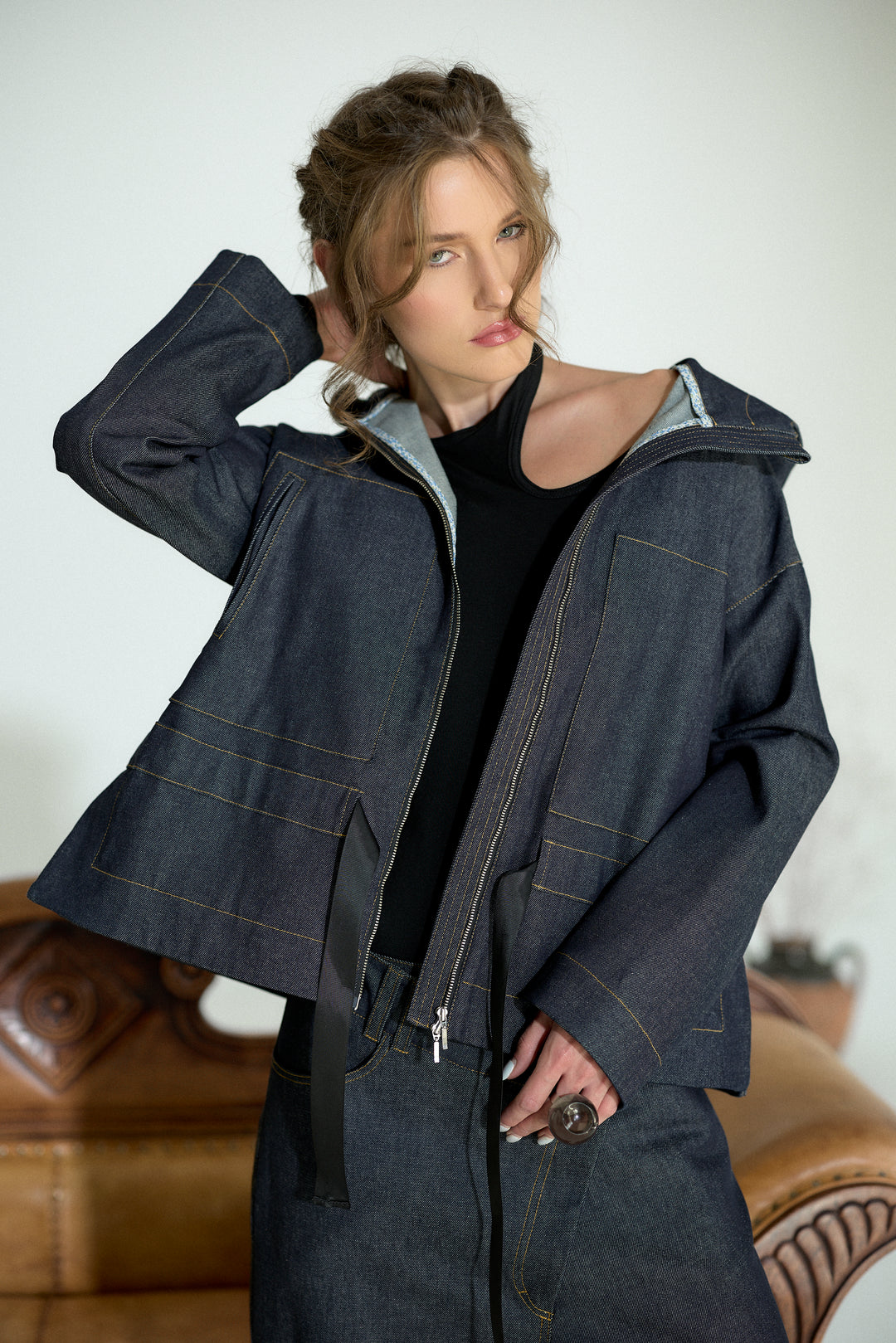 A woman models a dark indigo denim jacket with a zippered front, wide sleeves, and contrast gold-colored stitching, layered over a black top.