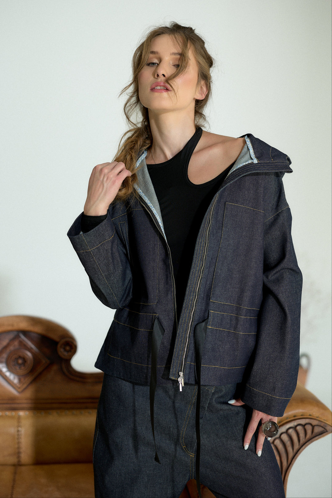 A woman wearing a dark indigo denim jacket with contrast gold stitching, a black top, and matching dark denim pants, posed in front of a light-colored wall with a brown leather sofa in the background.