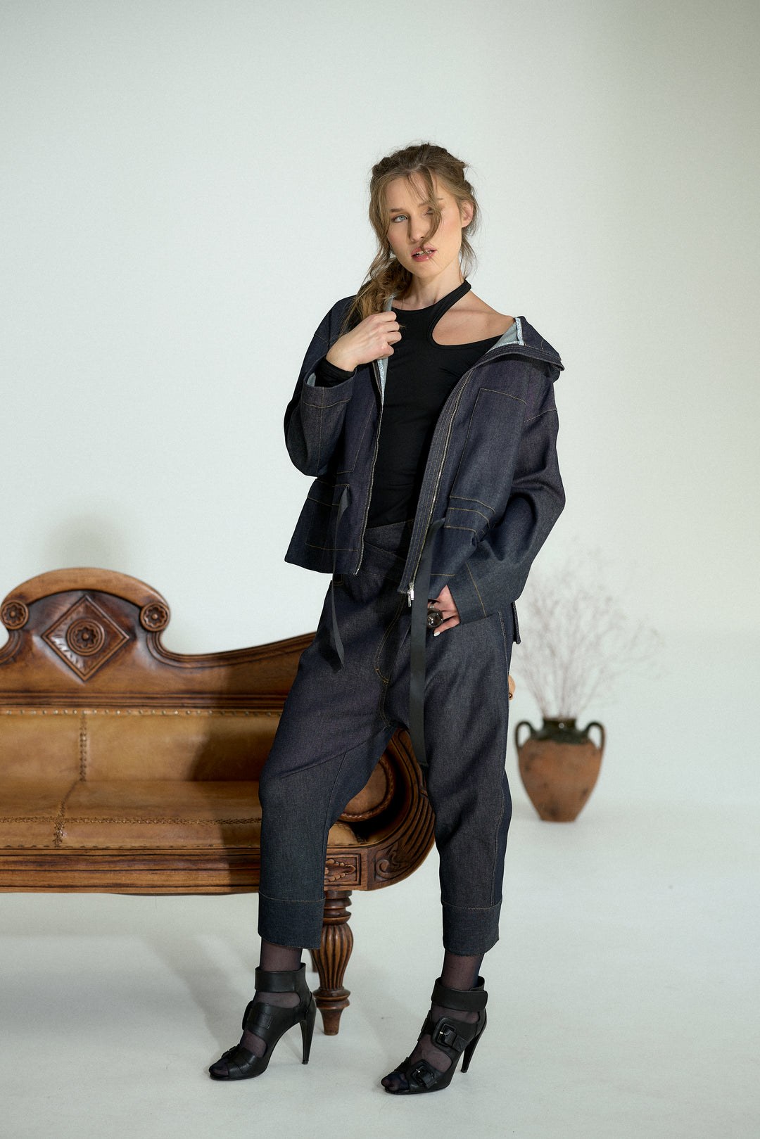 A woman models a dark washed denim two-piece set consisting of a cropped zip-up hoodie and matching relaxed-fit pants, styled with a black top and black high-heeled sandals, posed next to a vintage wooden bench.