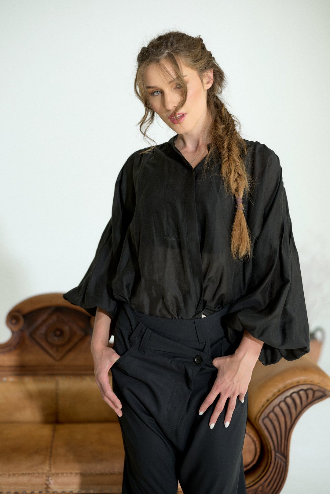 A woman wearing a black satin-like blouse with voluminous three-quarter sleeves and black trousers, standing in front of a light-colored wall with a brown leather sofa behind her.