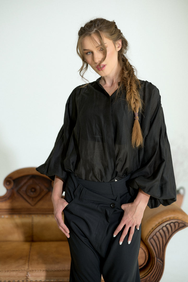 A woman wearing a black satin-like blouse with voluminous three-quarter sleeves and black trousers, standing in front of a light-colored wall with a brown leather sofa behind her.