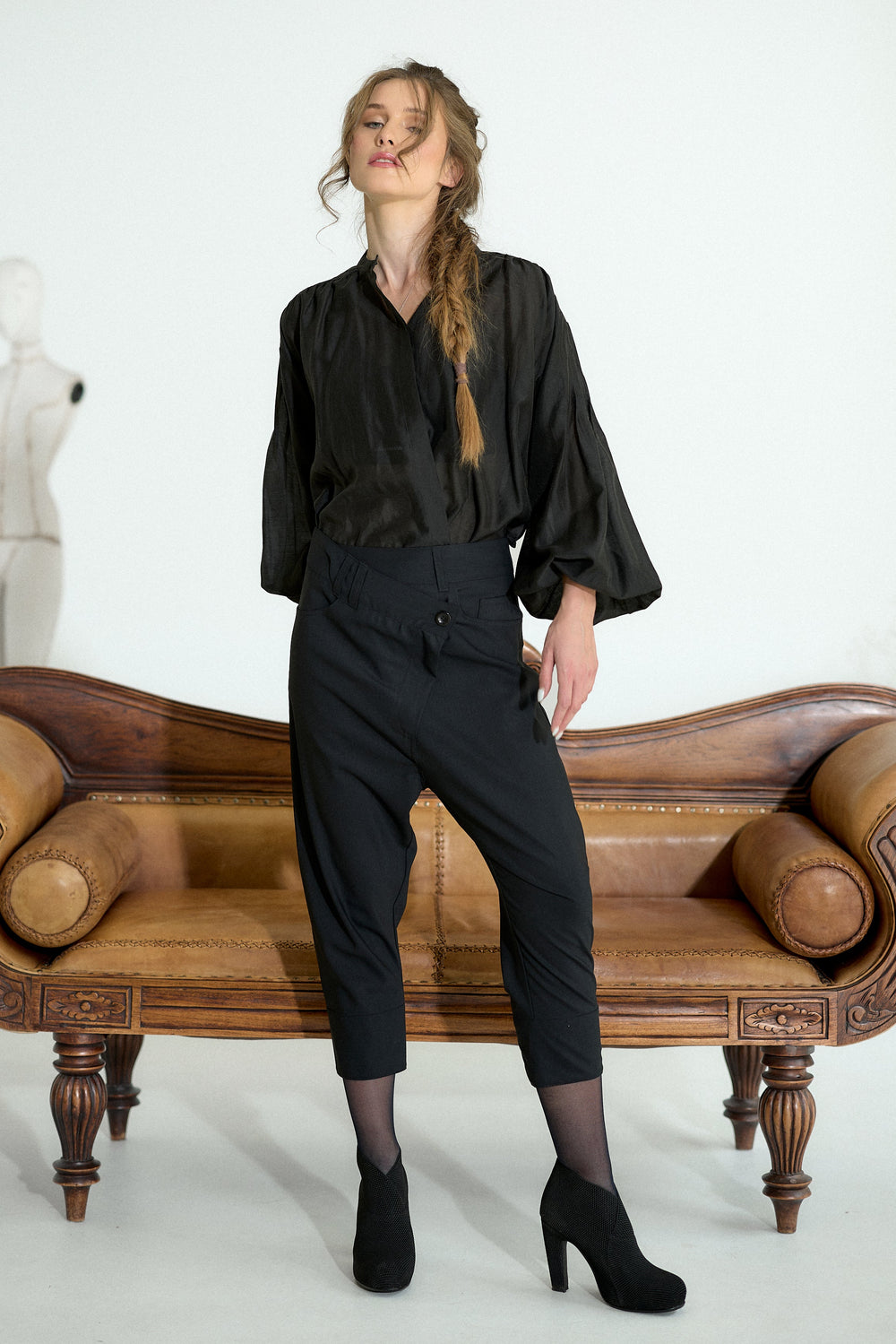 A woman models a black satin blouse and black cropped trousers with sheer black tights and black heels, standing in front of a brown leather bench against a white studio background.