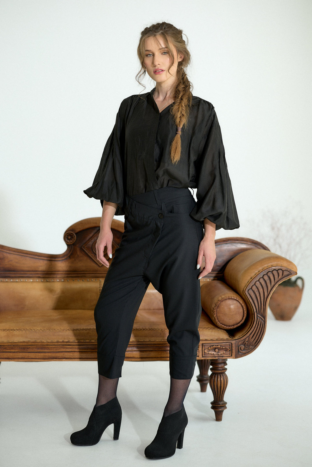 A woman models a black satin blouse with dramatic ruffled sleeves and black cropped trousers, standing in front of a vintage wooden chaise lounge against a white studio background.