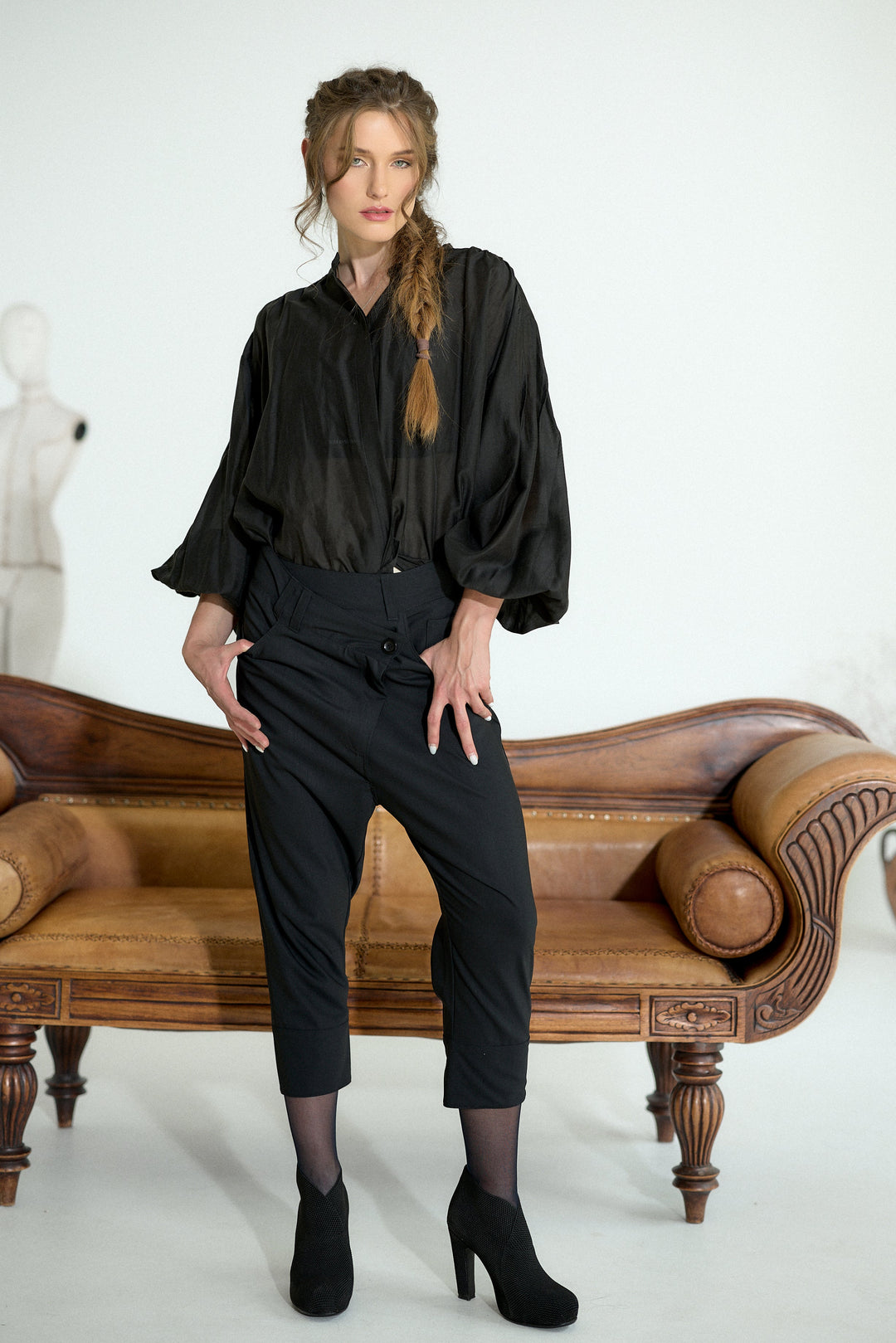 A woman models a black satin-like blouse with voluminous elbow-length sleeves and black cropped trousers, standing in front of a brown leather sofa against a white studio background.