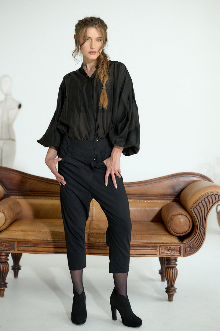 A woman models a black satin-like blouse with voluminous elbow-length sleeves and black cropped trousers, standing in front of a brown leather sofa against a white studio background.