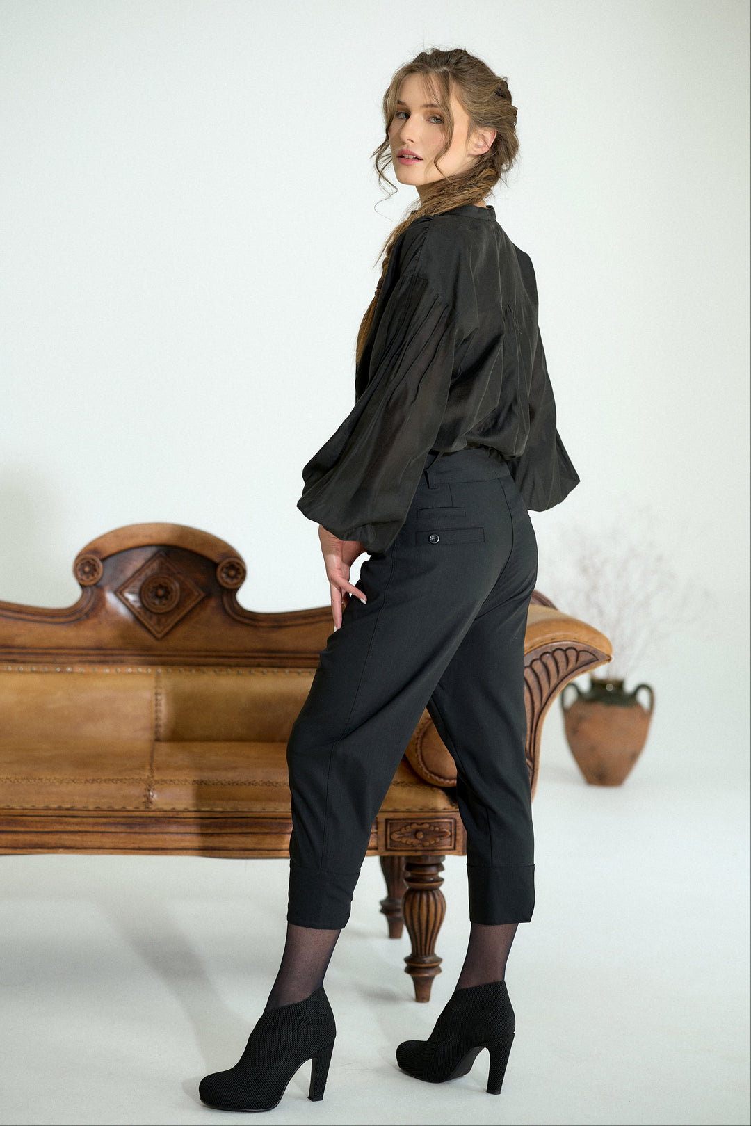 A woman posing in a black satin blouse and black cropped trousers, standing next to a vintage wooden bench in a studio setting.