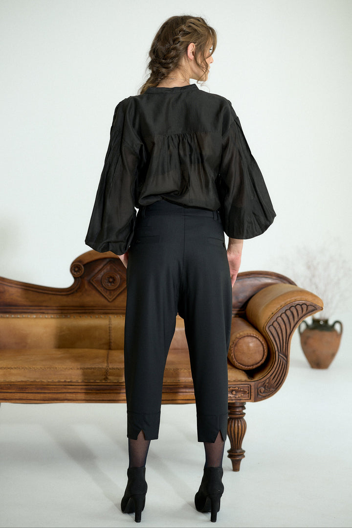 A woman stands with her back to the camera wearing a black sheer long-sleeve blouse with a gathered waist and black cropped trousers, posed in front of a brown leather chaise lounge against a white studio background.