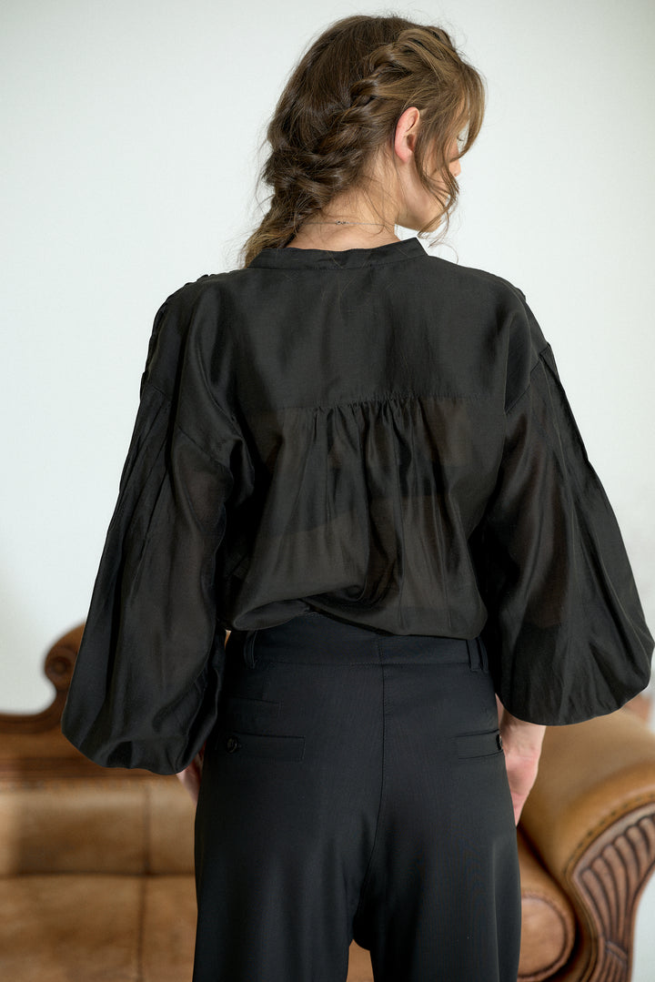 A woman viewed from behind wearing a black sheer long-sleeve blouse with a gathered back and black trousers, standing indoors near a brown leather chair against a light wall.