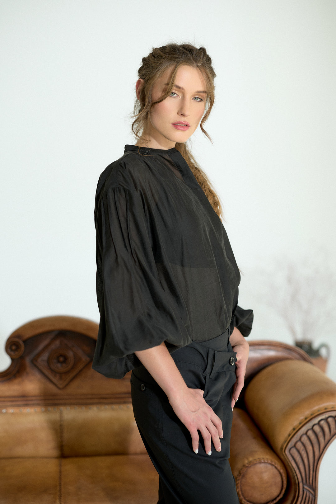 A woman wearing a black sheer long-sleeve blouse and black trousers, standing in front of a brown leather sofa against a white wall.
