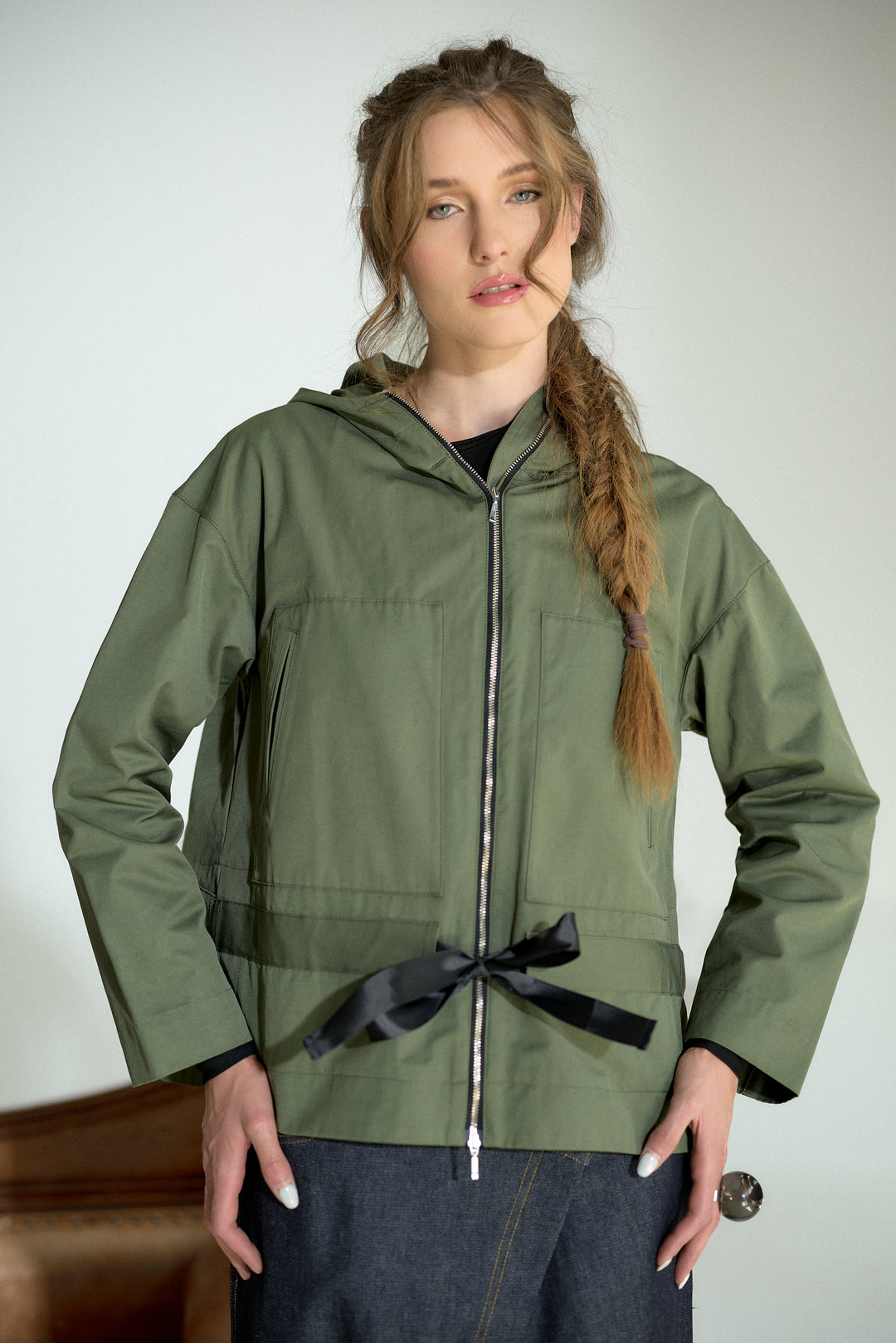 A woman modeling an olive green hooded jacket with a black ribbon tied at the waist, shown against a plain light gray background.