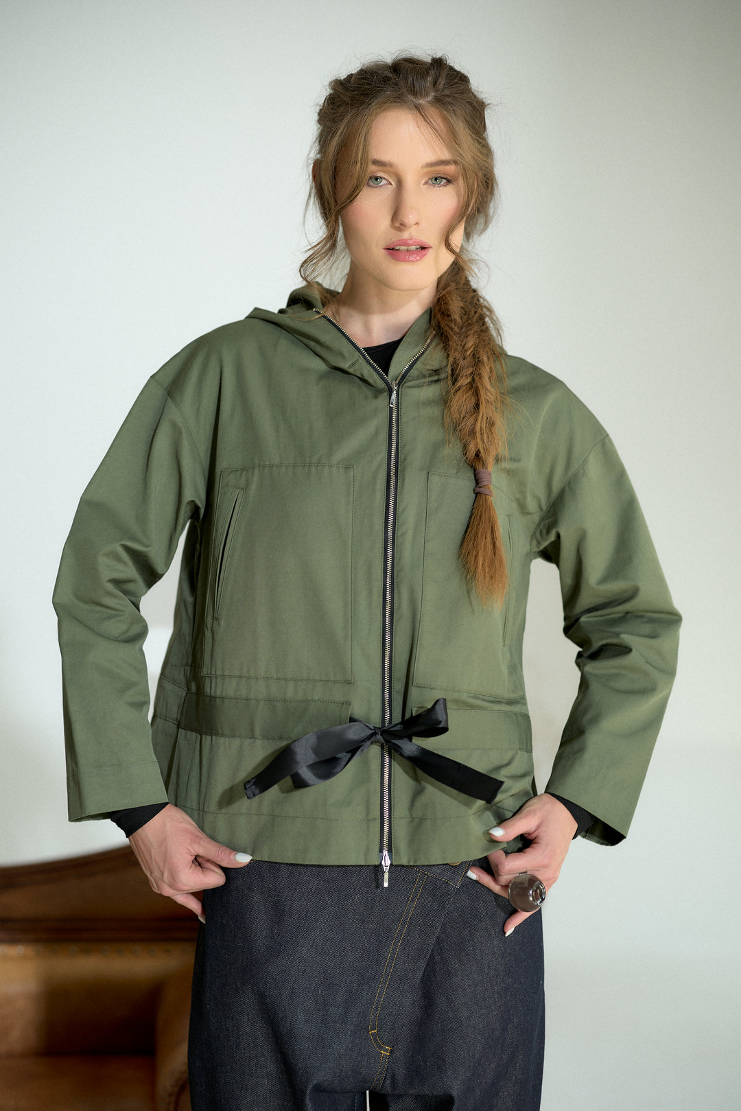 A woman modeling an olive green hooded jacket with a black front zipper, black waist tie, and black cuffs, paired with dark denim pants against a light gray background.