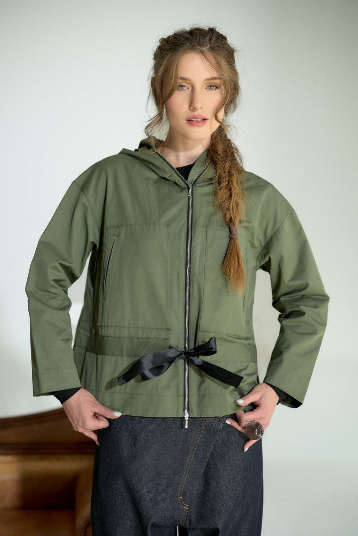 A woman modeling an olive green hooded jacket with a black front zipper, black waist tie, and black cuffs, paired with dark denim pants against a light gray background.