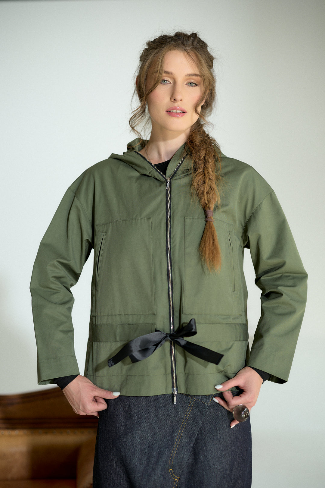 A woman modeling an olive green hooded jacket with a black ribbon tie at the waist, paired with dark denim jeans, against a plain light gray background.