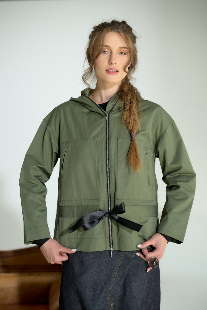 A woman modeling an olive green hooded jacket with a black ribbon tie at the waist, paired with dark denim jeans, against a plain light gray background.