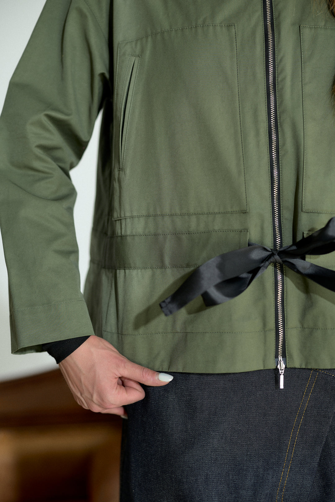An olive green utility jacket with a black satin tie belt at the waist, worn by a model with dark denim jeans.
