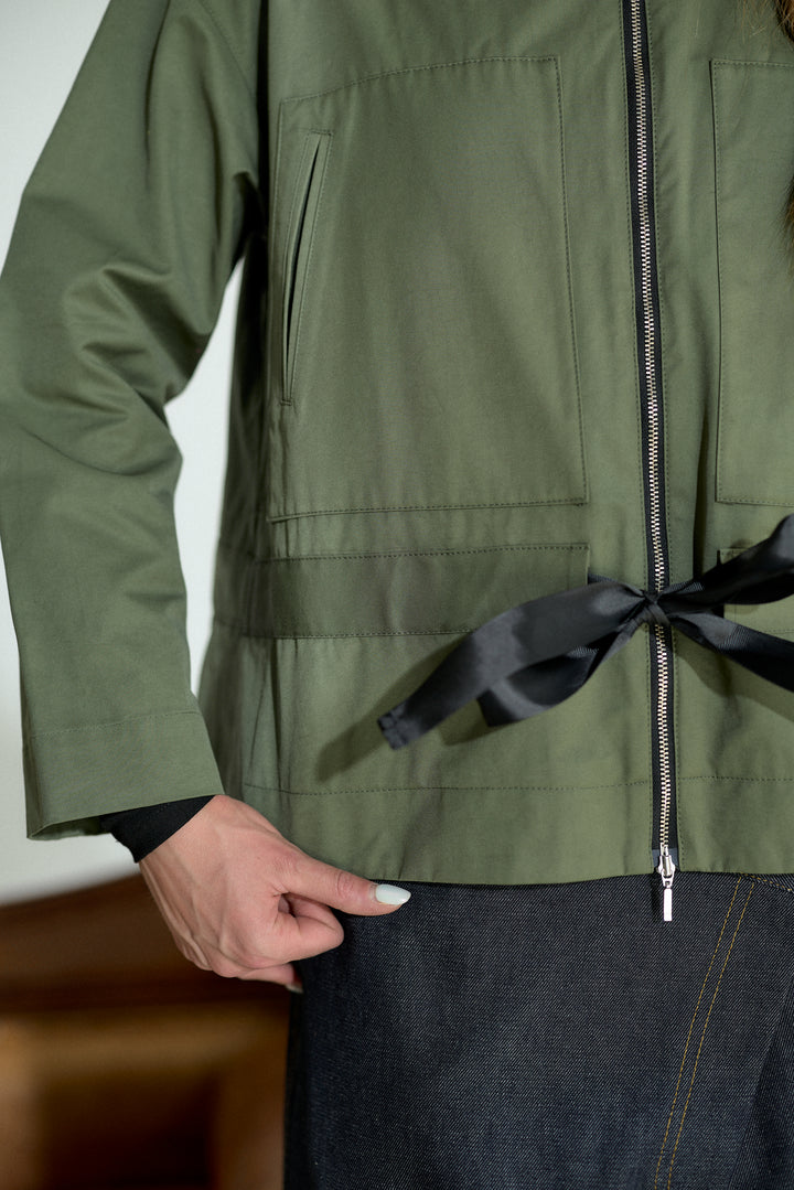 An olive green utility jacket with a black satin tie belt at the waist, worn by a model with dark denim jeans.