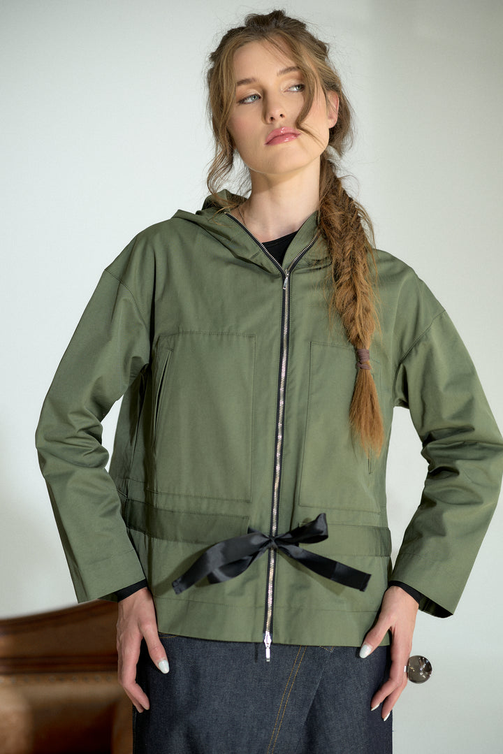 A woman wearing an olive green hooded jacket with a black ribbon bow at the waist, paired with dark gray pants, standing against a plain light background.