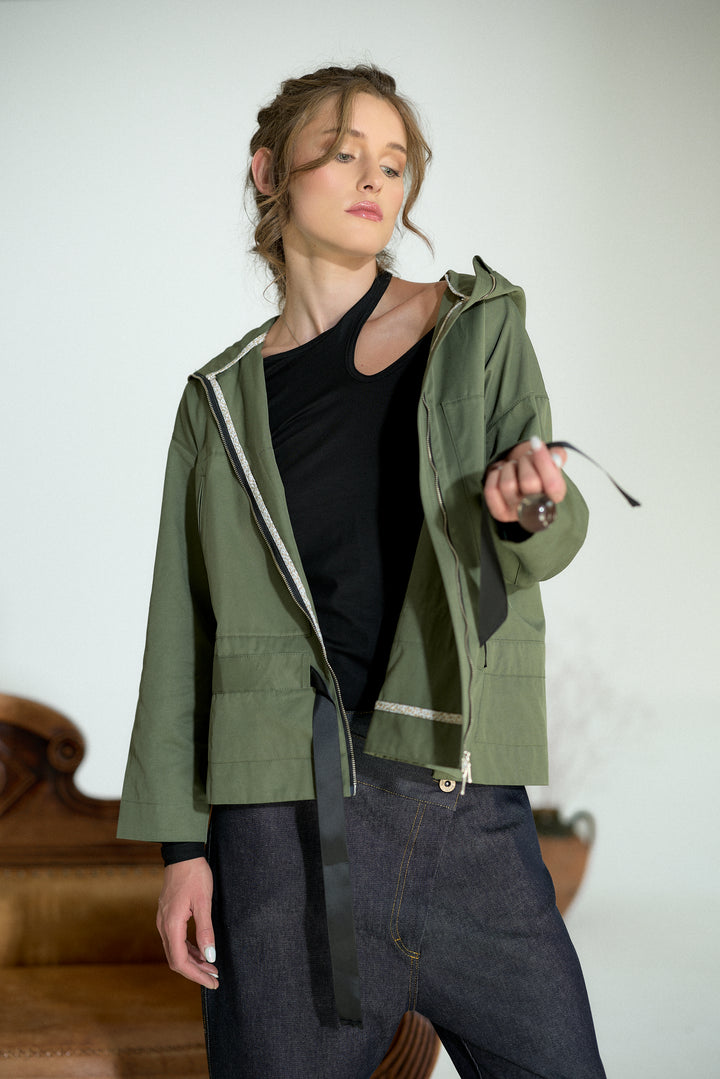 A woman models an olive green hooded jacket with a black zipper and a black waist tie, worn over a black top and dark jeans, set against a minimalist studio background.