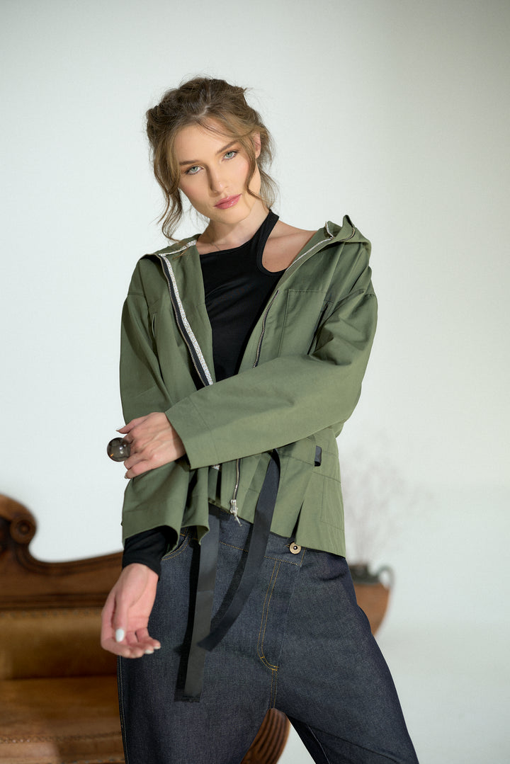 A woman modeling an olive green hooded jacket over a black top and dark jeans, posed in a minimalist indoor setting with a light gray wall and a wooden chair partially visible.