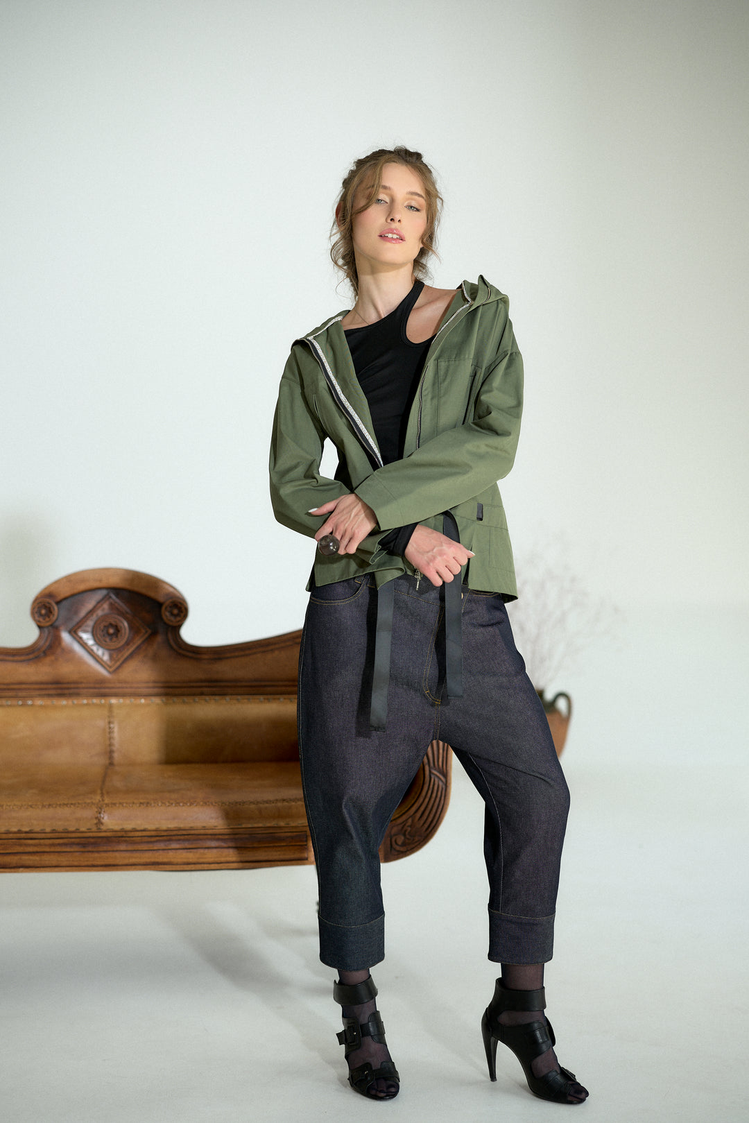 A woman models an olive green hooded jacket over a black top, paired with dark gray baggy pants and black ankle boots, standing in front of a white studio background with a wooden bench.