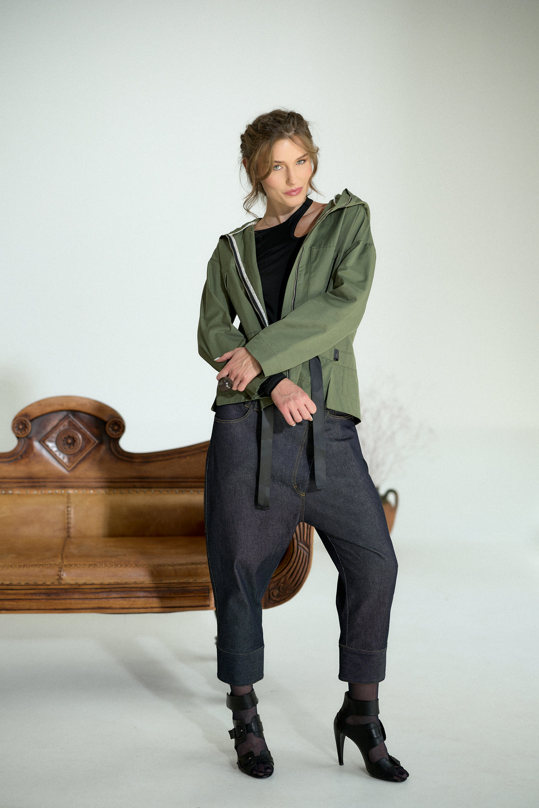 A woman models an olive green hooded jacket with a black top, dark gray baggy pants, and black high heels, standing in front of a white studio background with a wooden bench.