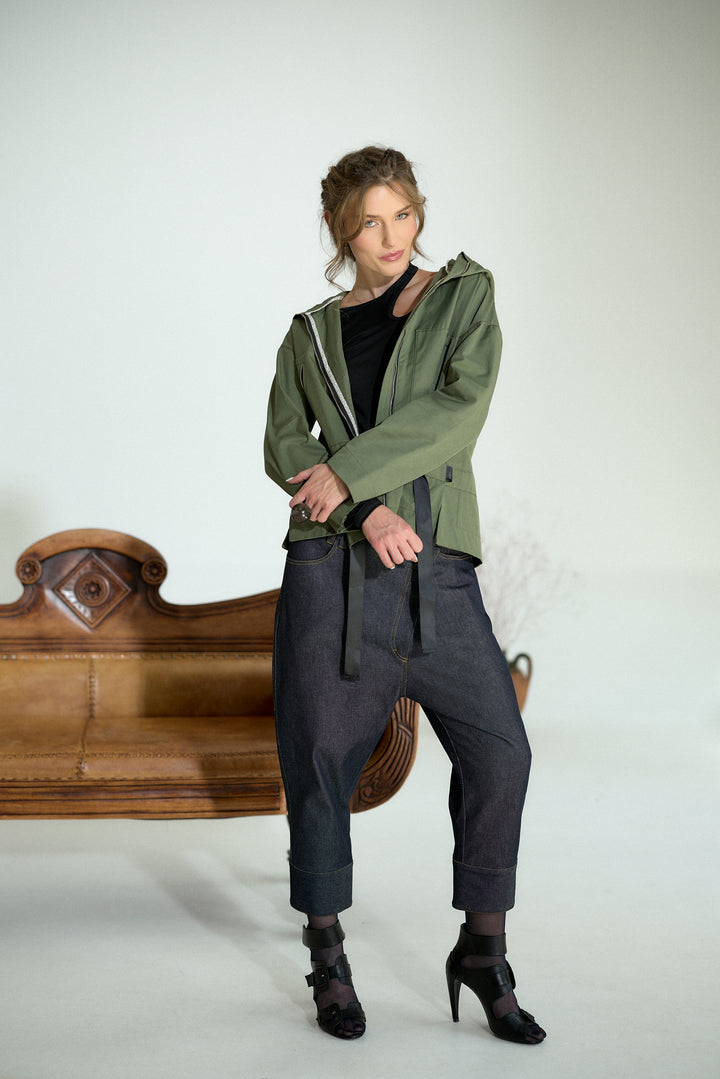 A woman models an olive green hooded jacket with a black top, dark gray baggy pants, and black high heels, standing in front of a white studio background with a wooden bench.