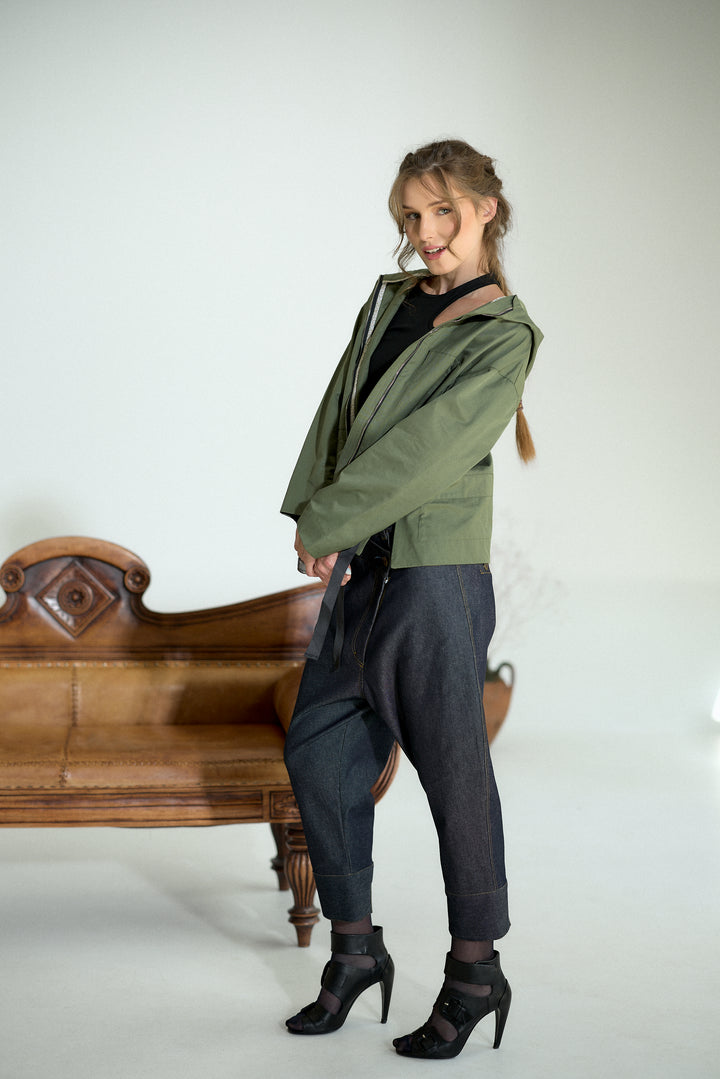 A woman models an olive green hooded jacket over a black top, paired with dark gray baggy pants and black high-heeled ankle boots, standing next to a vintage wooden bench against a white studio background.